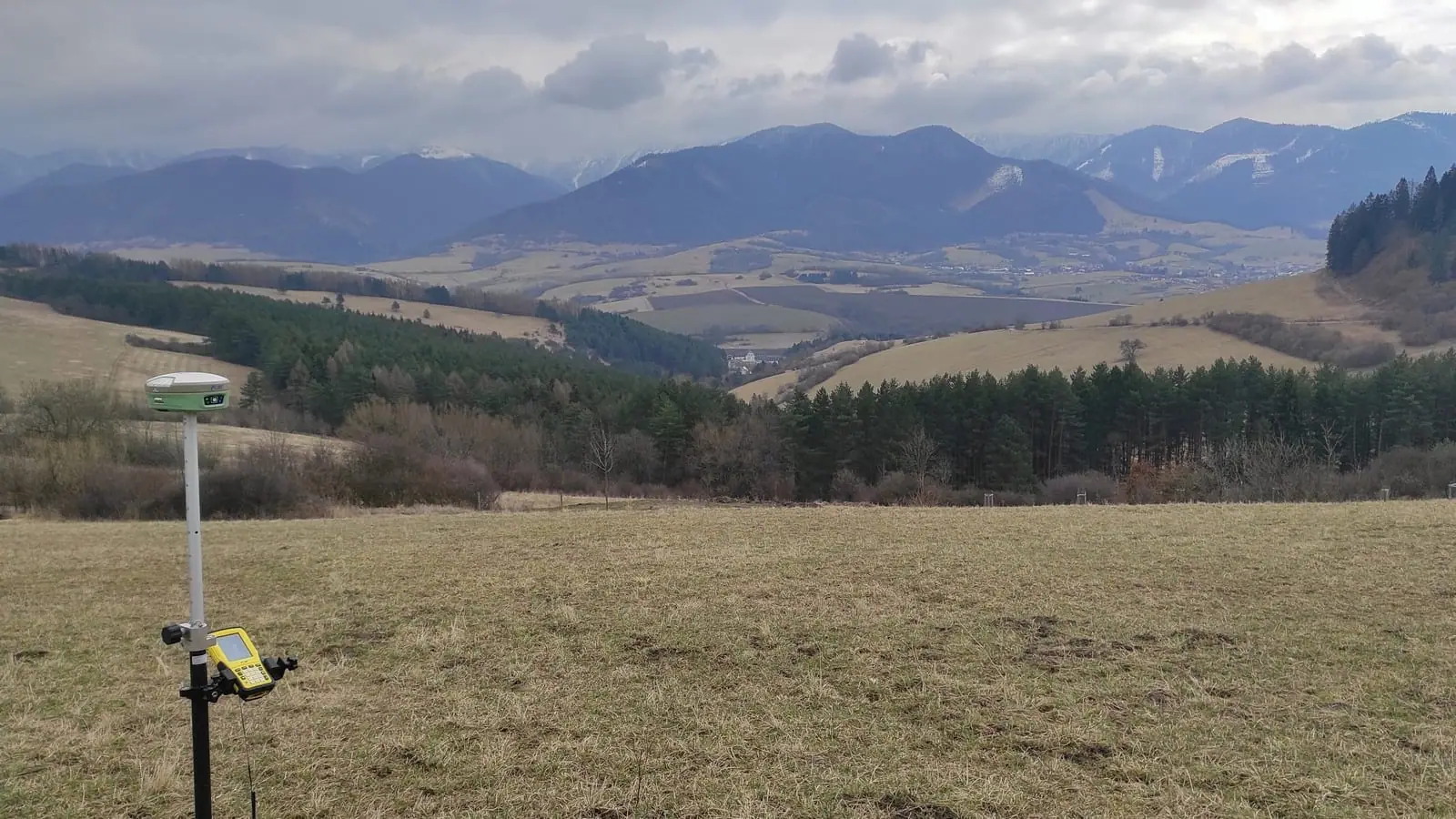 GNSS prijímač na výtyčke na lúke s výhľadom na zasnežené hory